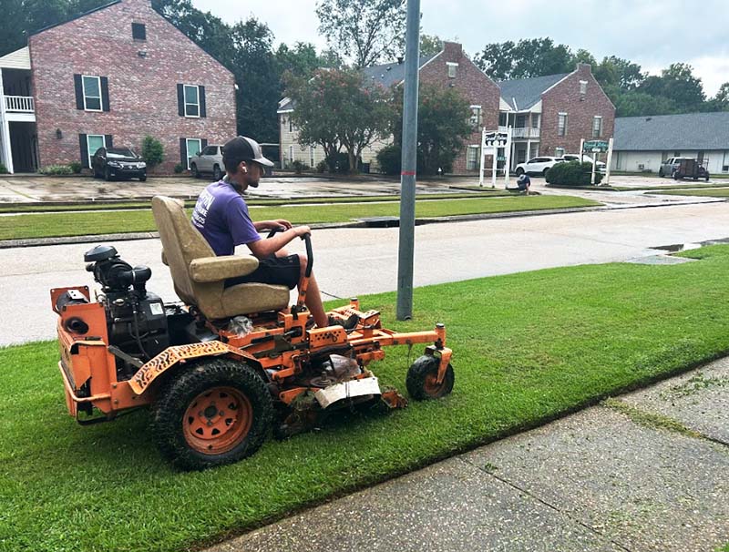 Person mowing lawn with riding mower on residential street.